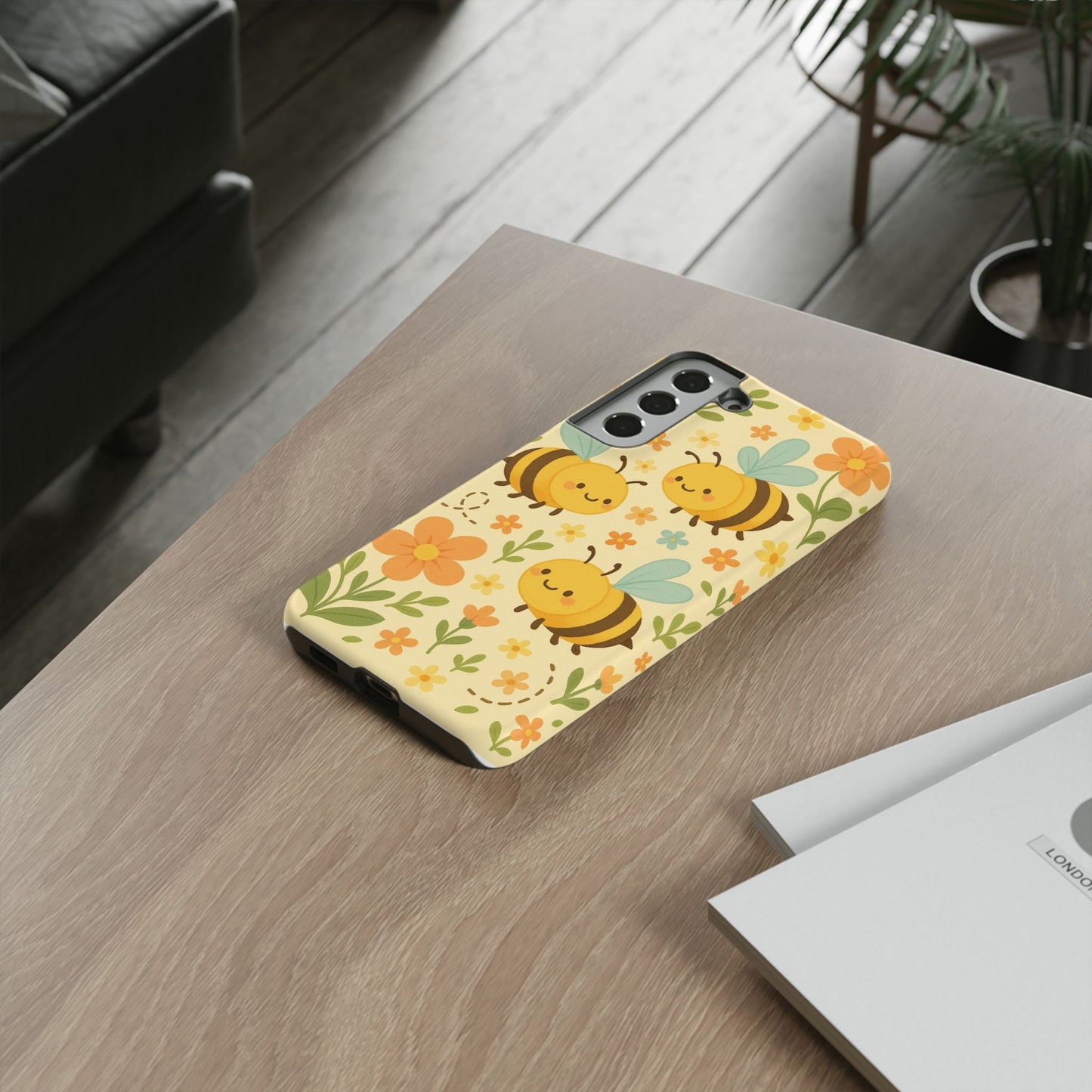 The Cute cartoon Floral Bee Tough Phone Case by Printify, with a yellow design of bees and orange flowers, rests on a wooden desk near a couch, plant, and papers—making it an ideal gift for bee lovers.