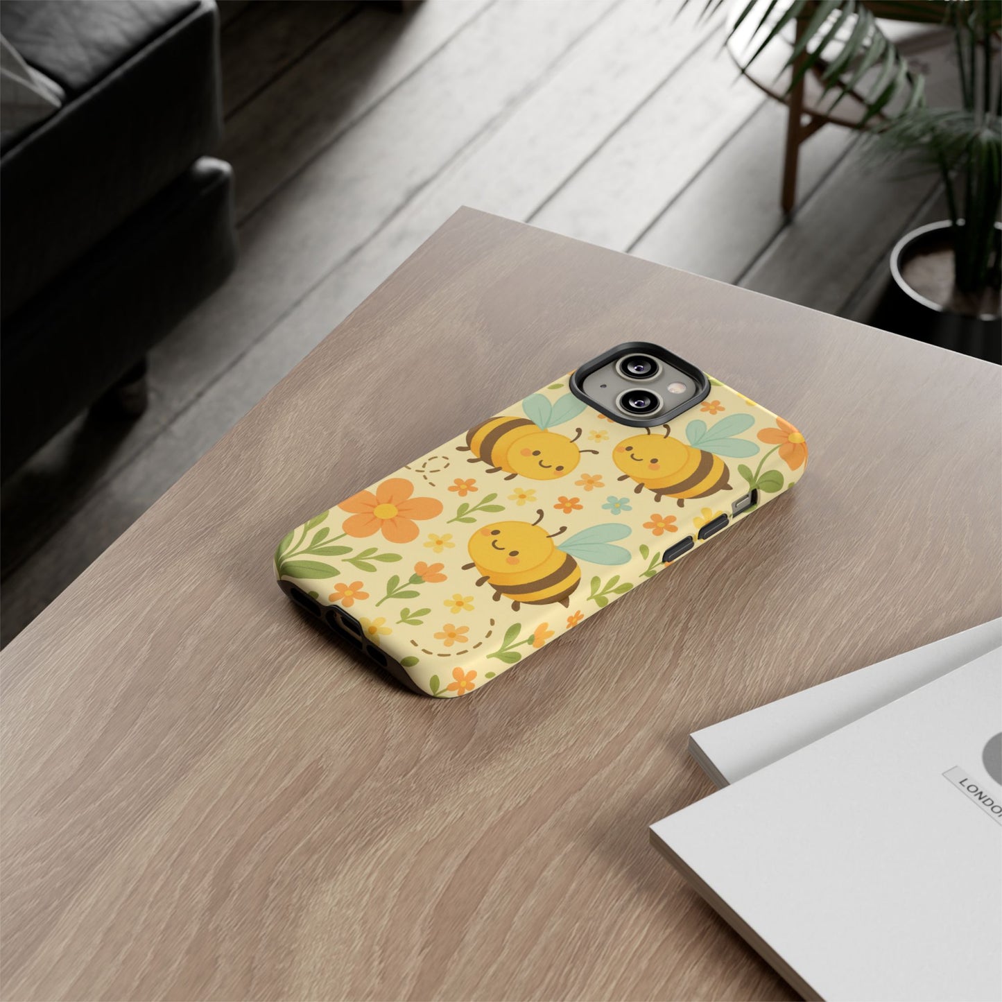A smartphone with the Cute cartoon Floral Bee Tough Phone Case by Printify, featuring smiling bees and flowers, rests on a wooden table beside a white envelope and paper—a perfect gift for bee lovers.