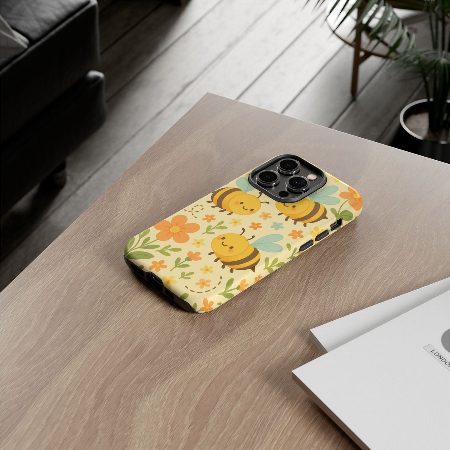 The Printify Cute Cartoon Floral Bee Tough Phone Case sits on a wooden table by white papers, with a plant and couch in the background. This stylish case is perfect for bee lovers, adding charm to your phone and any setting.
