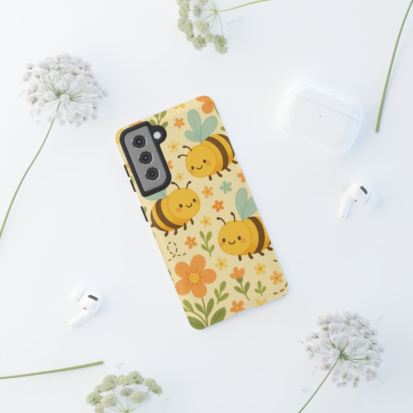 The Cute cartoon Floral Bee Tough Phone Case by Printify features a charming bee and flower design, displayed on a white surface with white flowers, wireless earbuds, and a charging case—an ideal gift for bee lovers.