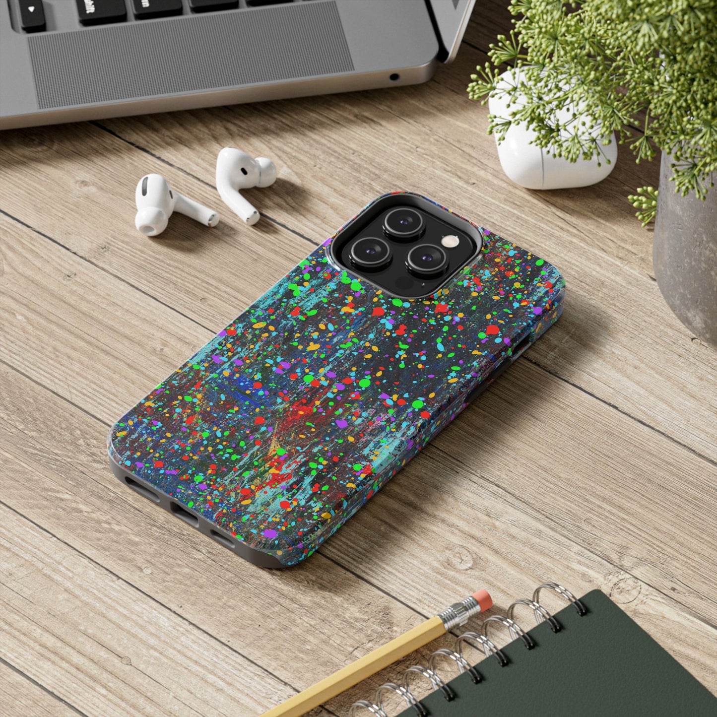 Printify colorful abstract drip paint phone case on wooden desk beside AirPods laptop plant and notebook