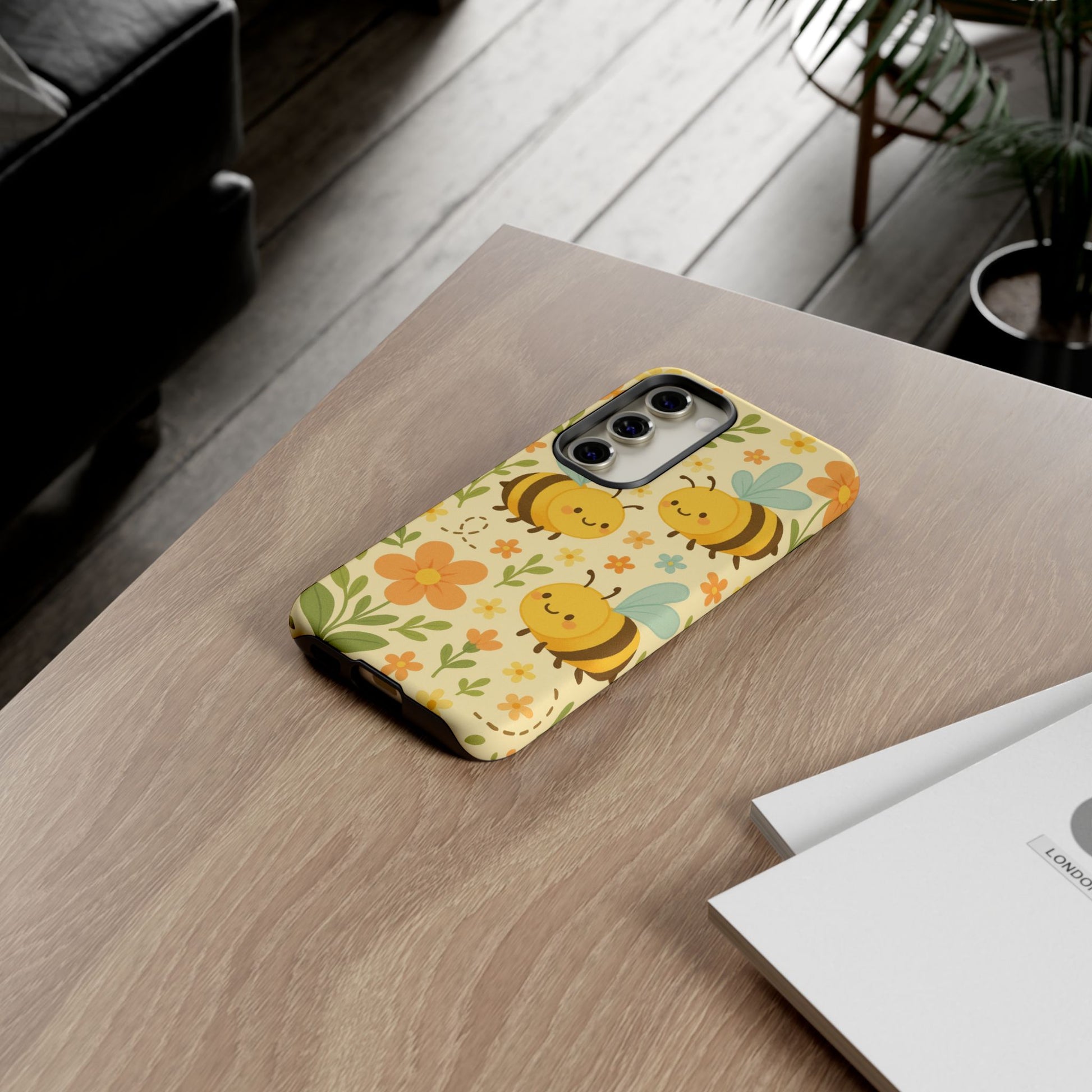 The Cute cartoon Floral Bee Tough Phone Case by Printify, featuring smiling bees, flowers, and leaves, sits on a wooden table with white papers in a cozy room with plants and a sofa—an ideal bee lovers gift!.