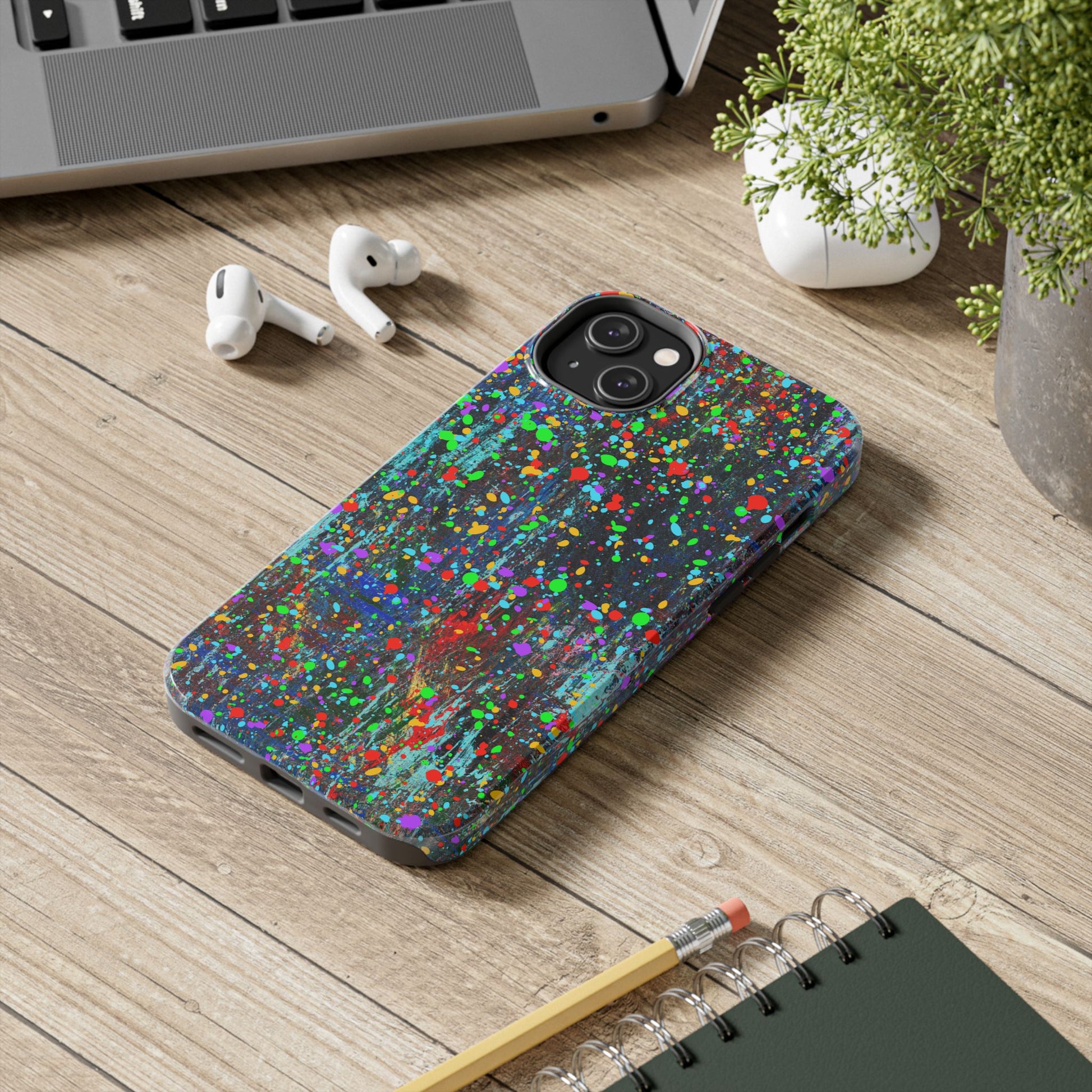 Printify colorful abstract drip paint phone case on wooden desk beside laptop earbuds and notebook